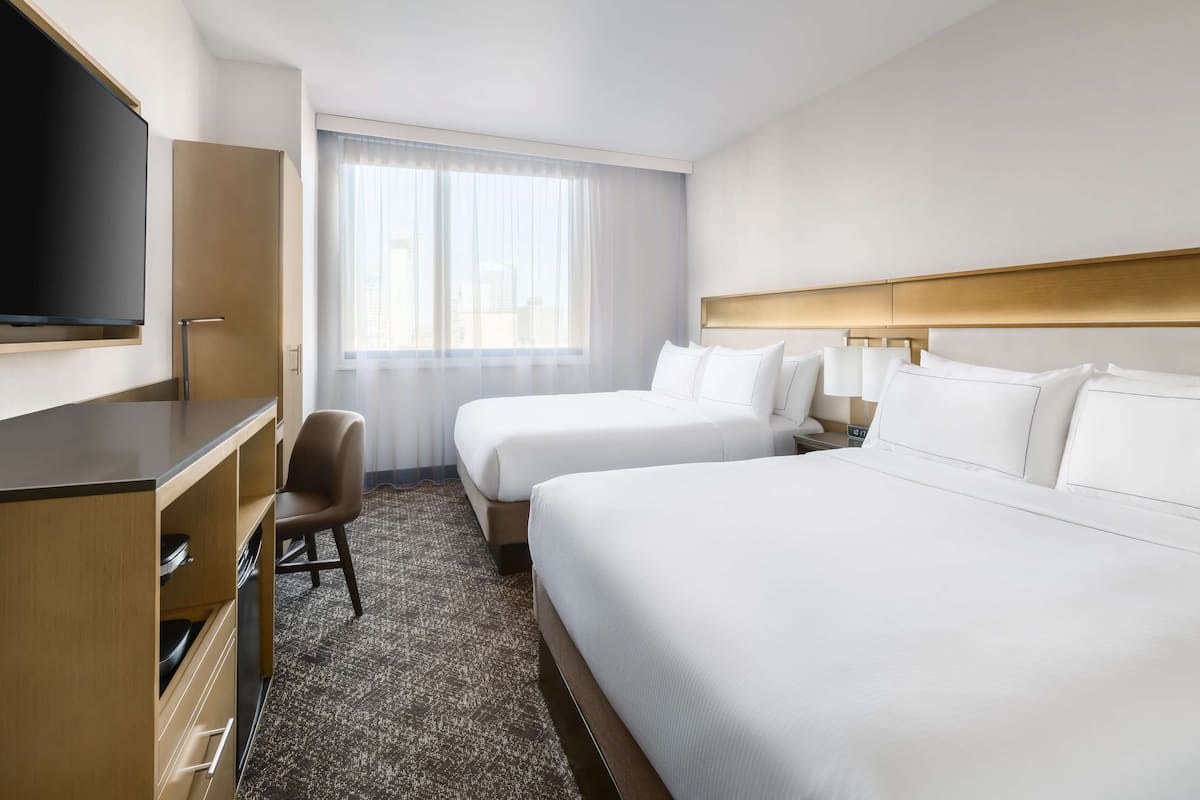 Ein Hotelzimmer mit zwei Betten, einem Schreibtisch, einem Sessel, einem Fernseher und einem großen Fenster mit Vorhängen.