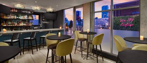 A bar area with a city view, tables, chairs, and a variety of bottles on shelves.