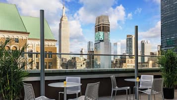Terraço no telhado com cadeiras e mesa de metal, com vista para um horizonte urbano com diversos edifícios.