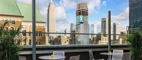 A rooftop terrace with metal chairs and a table, overlooking a cityscape with various buildings.