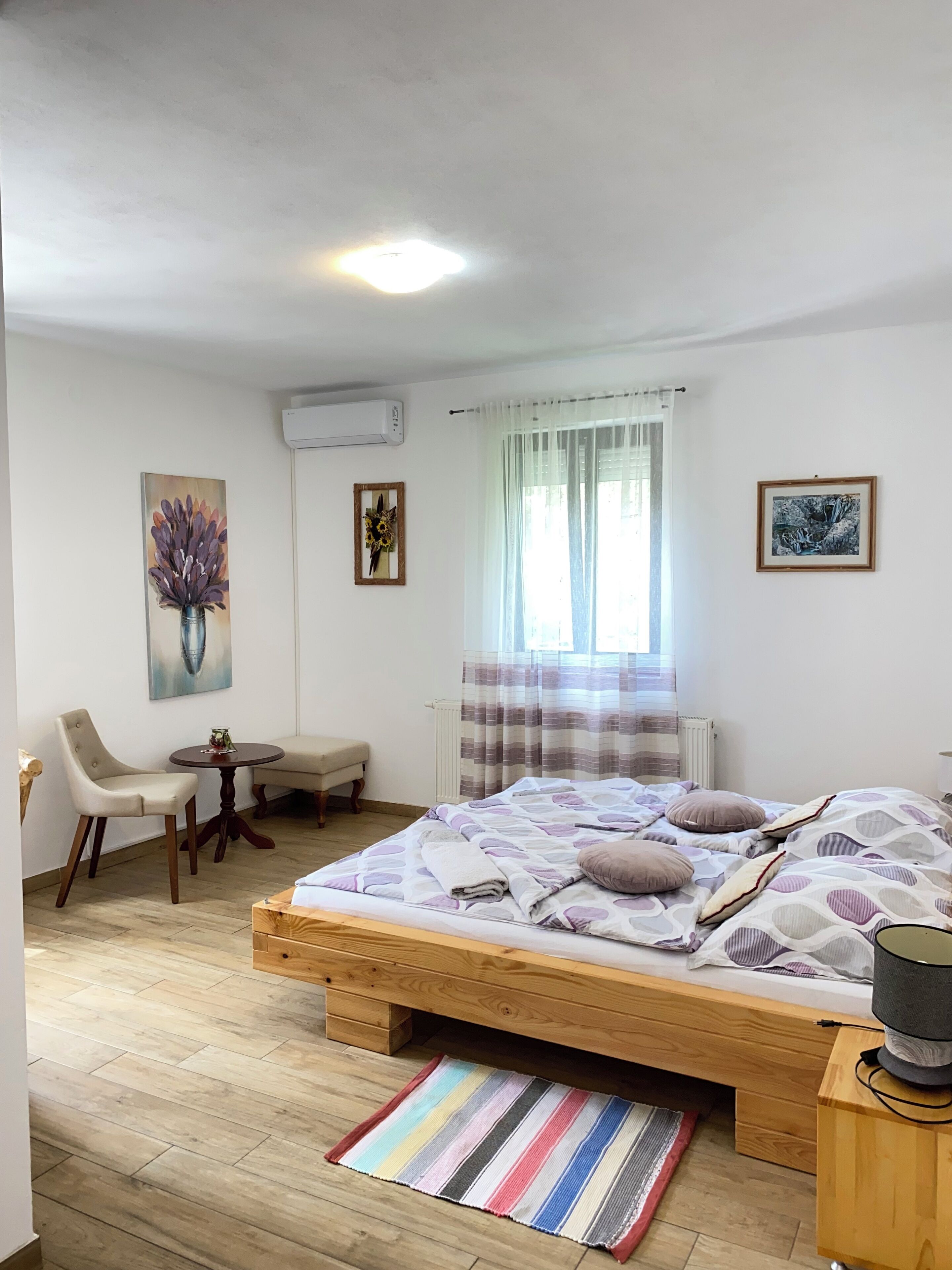 A bedroom with a wooden bed, a bedside table, a chair, a small round table, and a window with curtains.
