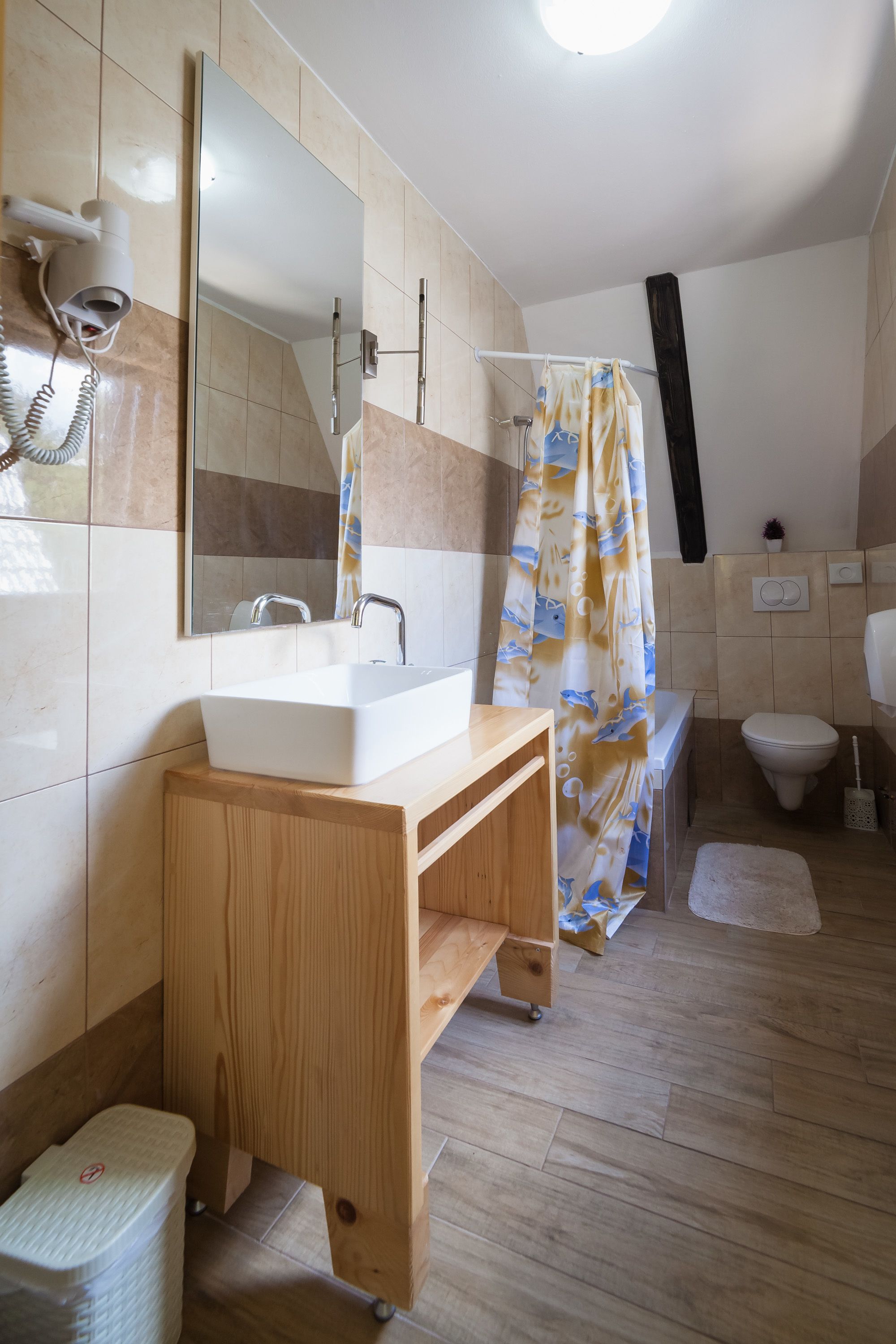 Un bagno con un mobile contenitore in legno, un lavandino bianco, uno specchio, una tenda da doccia, un water e un soffione doccia manuale.