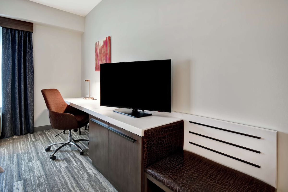 A hotel room with a desk, chair, television, and a bench.
