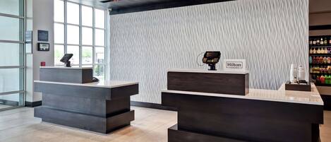 A modern hotel reception area with two dark reception desks, a textured wall, and a visible Hilton Honors sign.