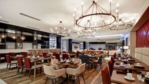 Ein modernes Restaurant mit verschiedenen Essbereichen, darunter runde Tische und Boxsitzgruppen, sowie einer zentralen Bar.