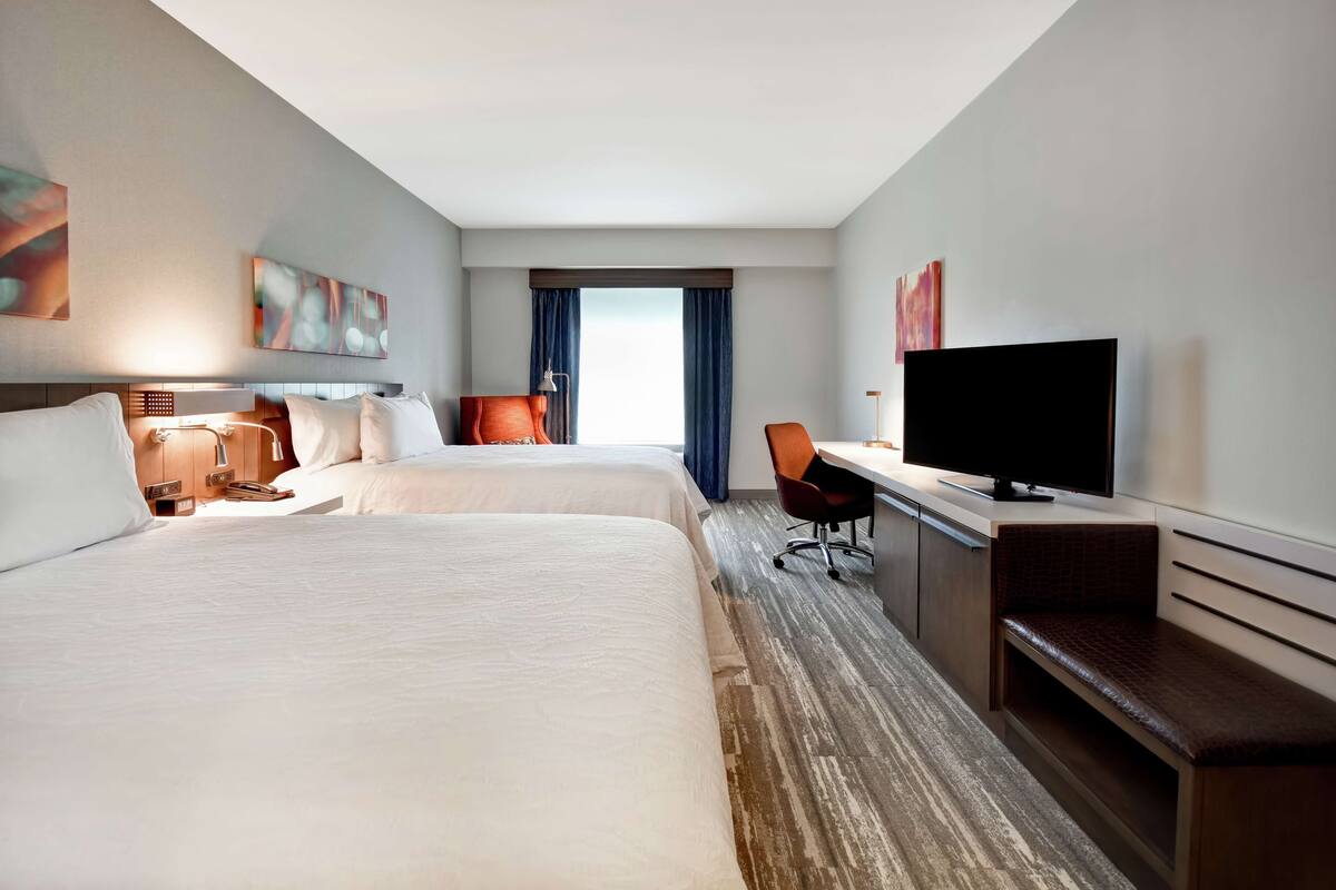A hotel room with two beds, a desk, a television, and a window with curtains.