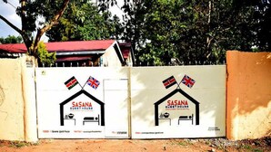 A gated entrance with two identical signs for 'SASANA GUEST HOUSE' featuring flags of Kenya and the UK, and contact information.