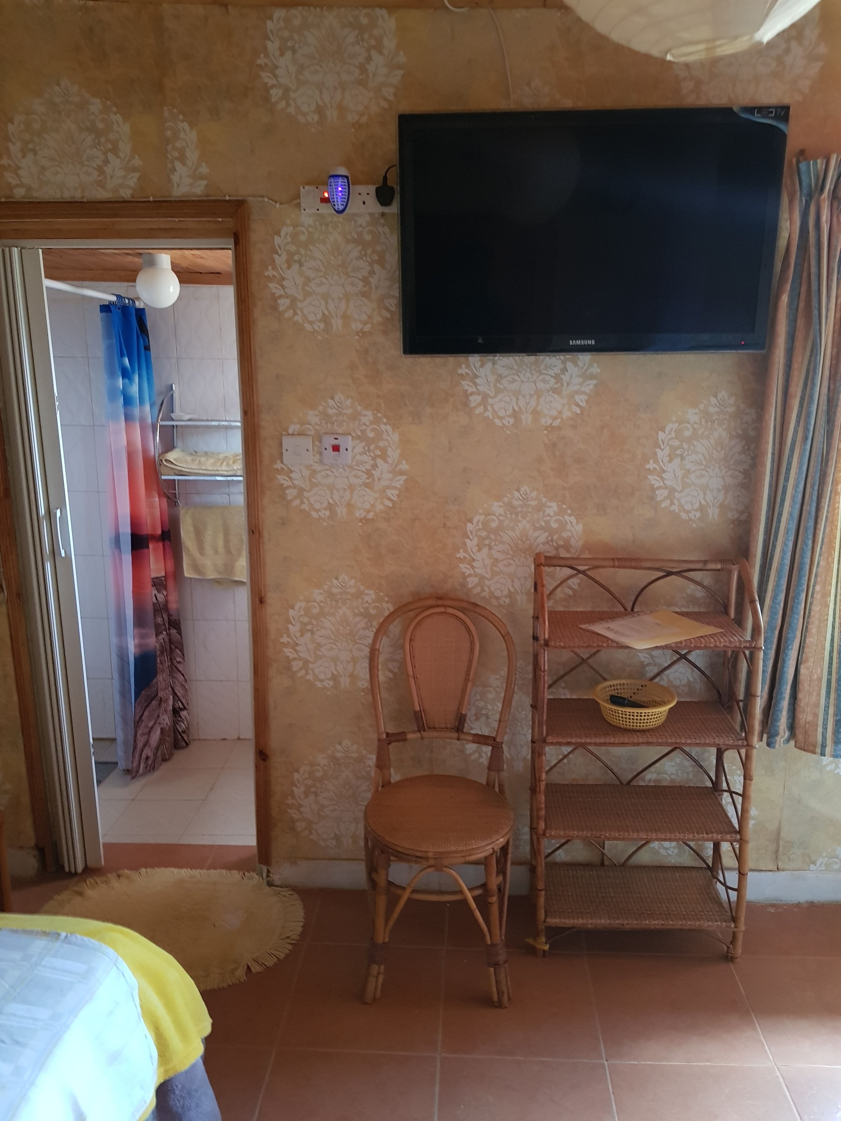 A room with a television mounted on the wall, a wicker chair, a shelf with items, and a door leading to another room.