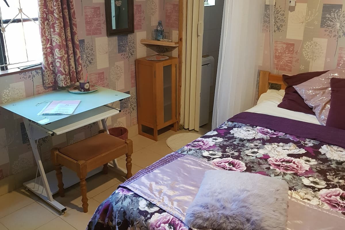 Ett sovrum med en säng, ett skrivbord och en stol. Det finns ett fönster med gardiner, en spegel och en hylla med dekorativa föremål.