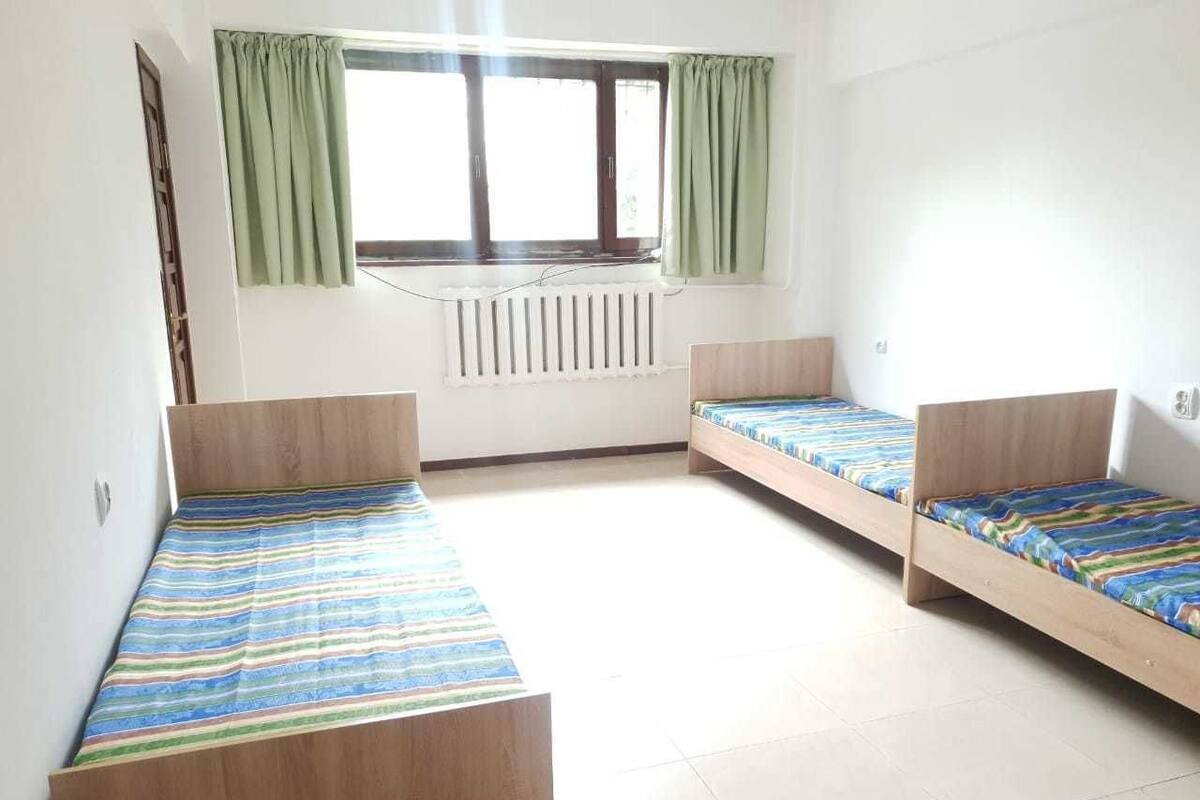 A dormitory room with two single beds, a radiator, a window with curtains, and a radiator above the window.