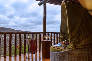 Una terraza de madera con una silla de mimbre y una mesa dispuesta con una copa de vino, una botella y un cuenco de fruta.