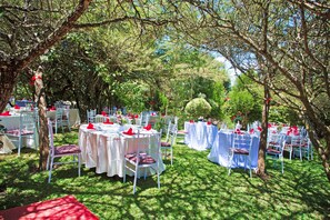 An outdoor event space with round tables and white chairs, surrounded by trees.