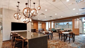 A modern hotel lobby with a central bar area, multiple seating arrangements, and large windows providing natural light.
