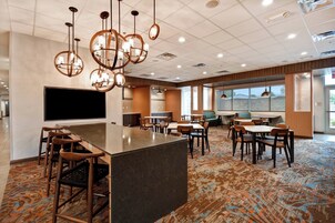 Una moderna hall d'albergo con un'area bar centrale, diverse disposizioni di posti a sedere e ampie finestre che lasciano entrare la luce naturale.