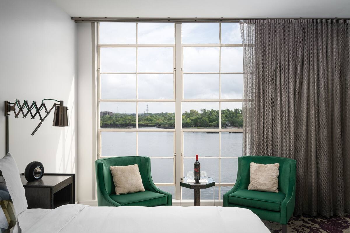 A hotel room with a large window, two green armchairs, a small table with a bottle and glasses, and a bed with a white comforter.