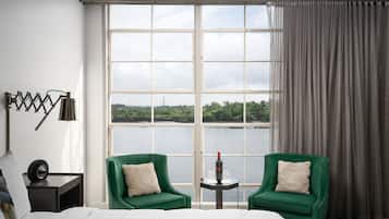 Een hotelkamer met een groot raam, twee groene fauteuils, een kleine tafel met een fles en glazen, en een bed met een witte deken.