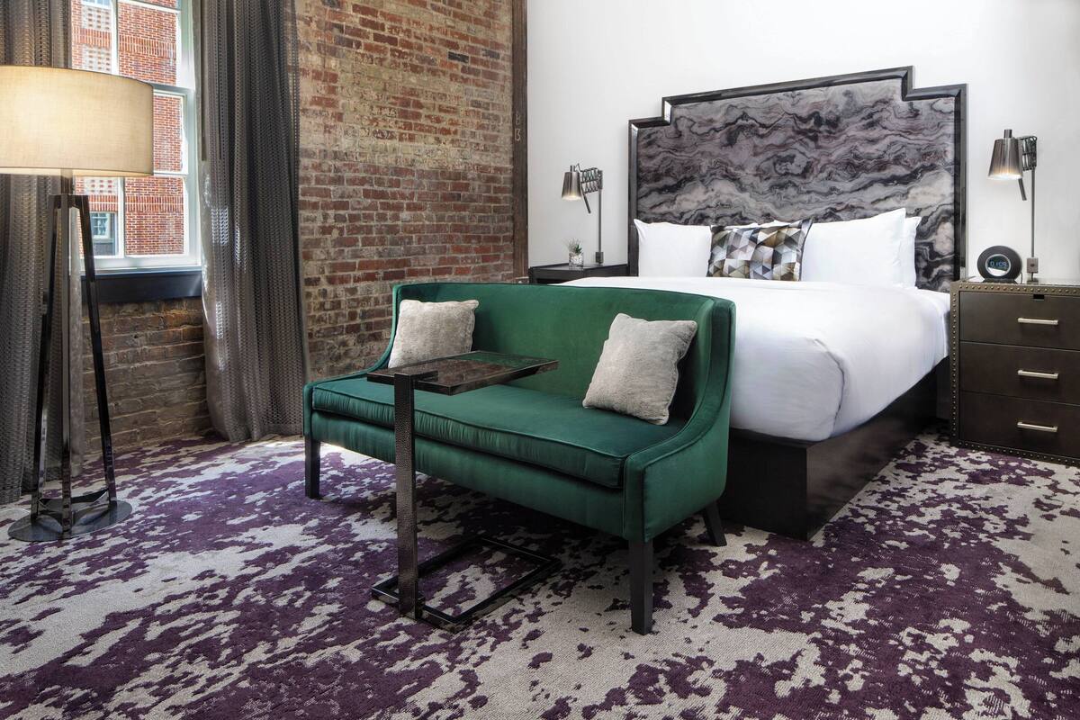 A hotel room with a green sofa, a large bed with a marble headboard, a brick wall, and a purple patterned carpet.