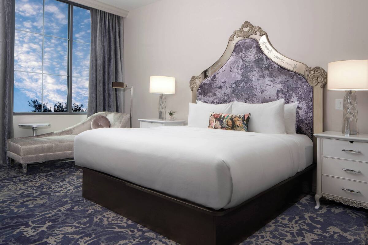 A hotel room with a large bed, a chaise lounge, a nightstand with a lamp, and a view of the sky through a window.