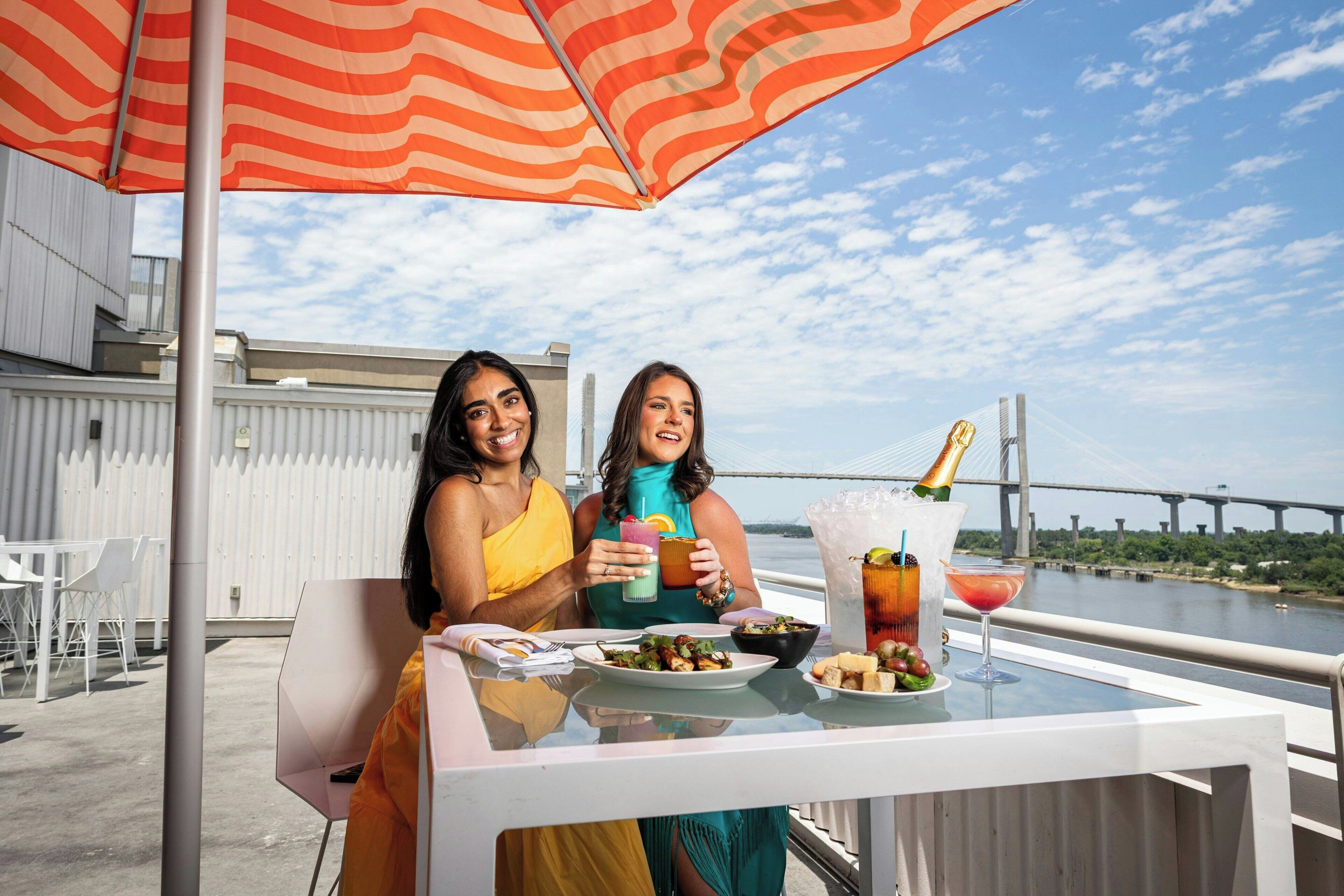 Two people sitting at a table with drinks and food, overlooking a bridge and water.