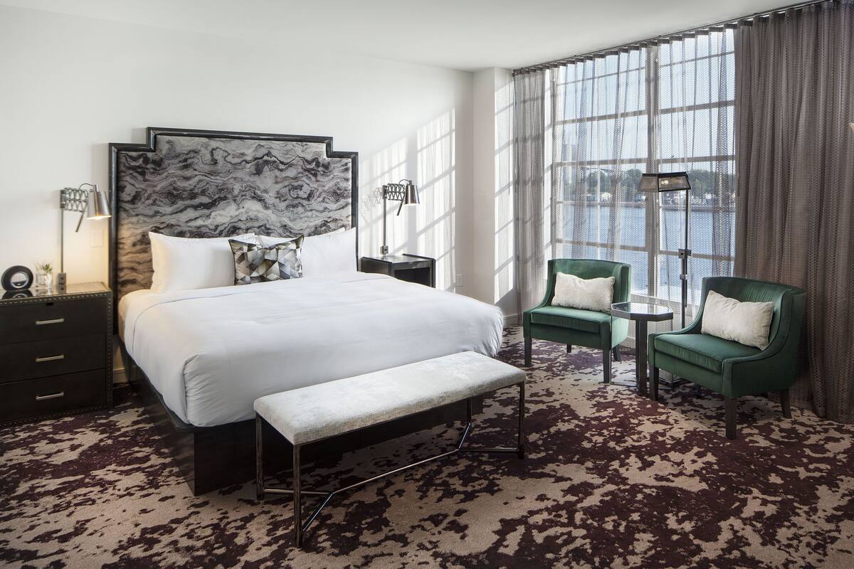 A hotel room with a large bed, two chairs, a bench, a nightstand, a lamp, and a view of a body of water through large windows.