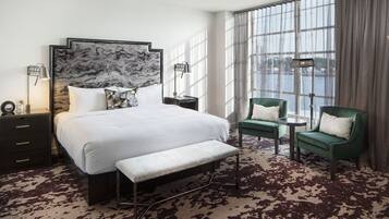 Une chambre d’hôtel avec un grand lit, deux fauteuils, un banc, une table de chevet, une lampe et une vue sur une étendue d’eau grâce à de grandes fenêtres.