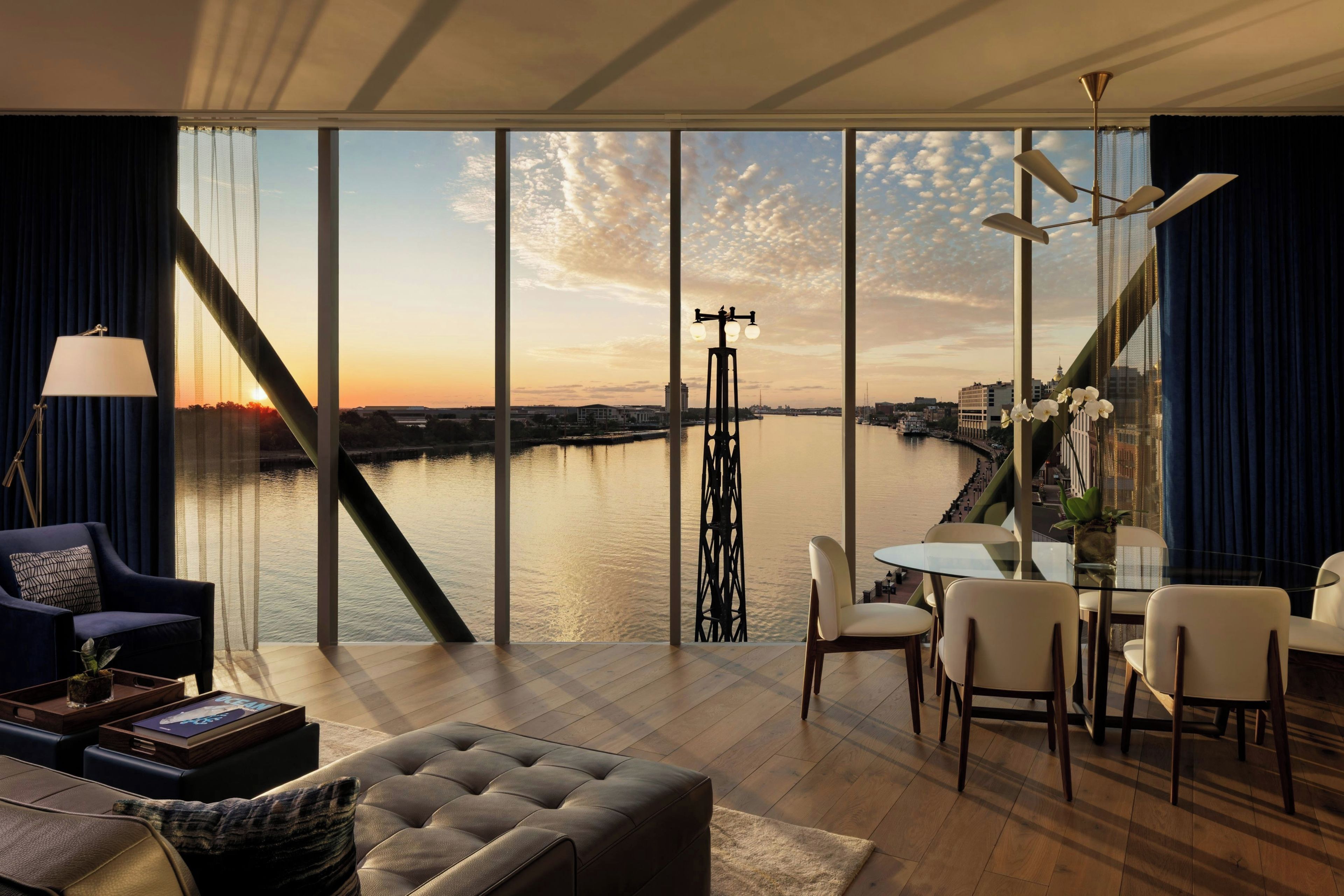 A modern living room with a large window overlooking a river, a dining area with a glass table, and a sofa with a decorative pillow.
