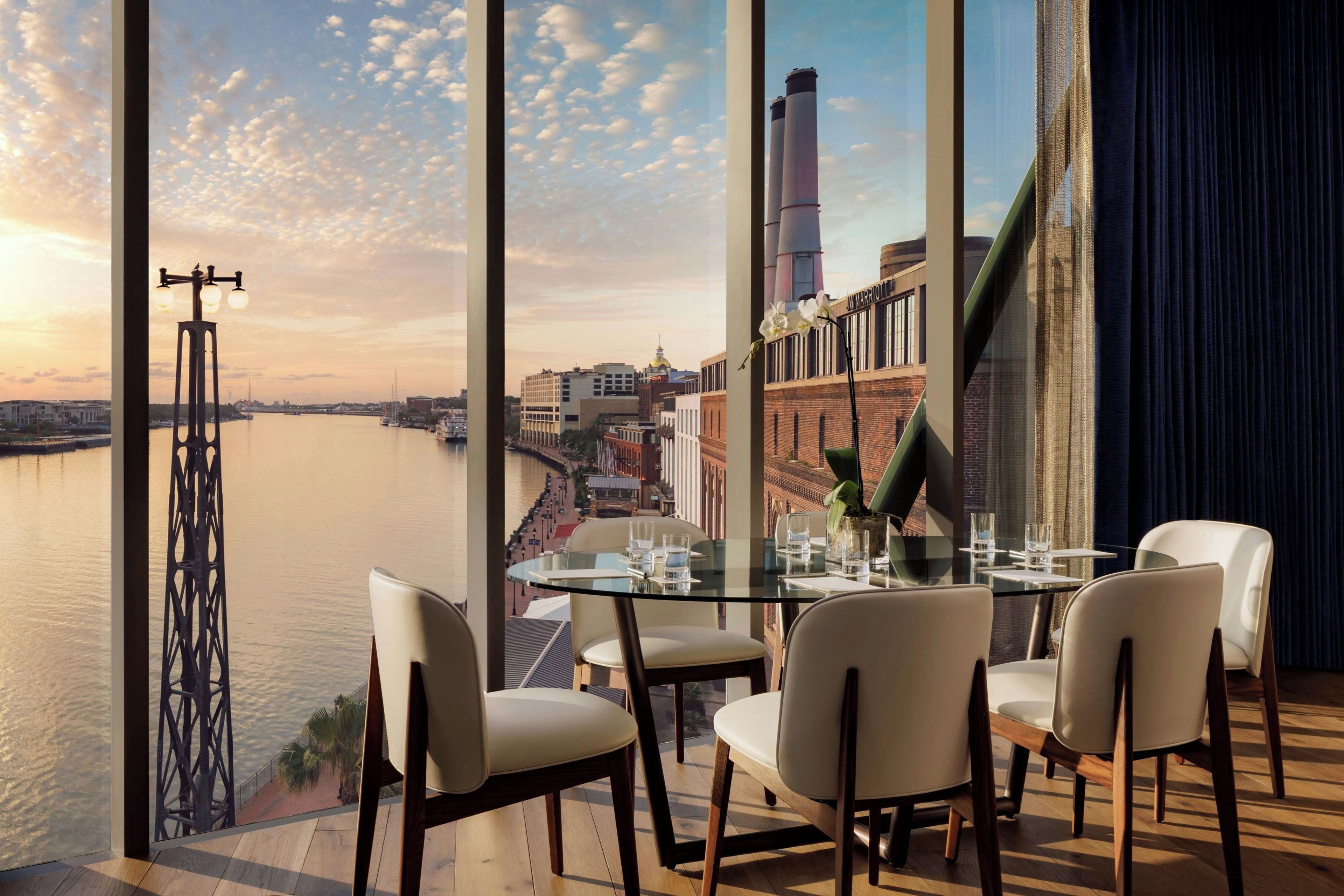 A dining area with a view of a river and cityscape, featuring a table set for four, chairs, and a large window.