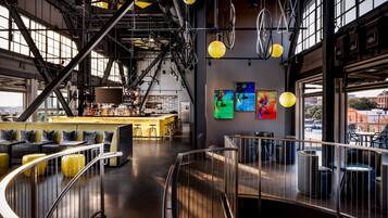 A modern bar area with a high ceiling, exposed beams, and a bar counter. There are multiple seating areas with stools and round tables.