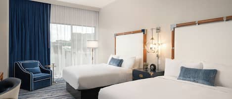 Habitación de hotel con dos camas, una silla, una mesita de noche, una lámpara y vista a la ciudad por la ventana.