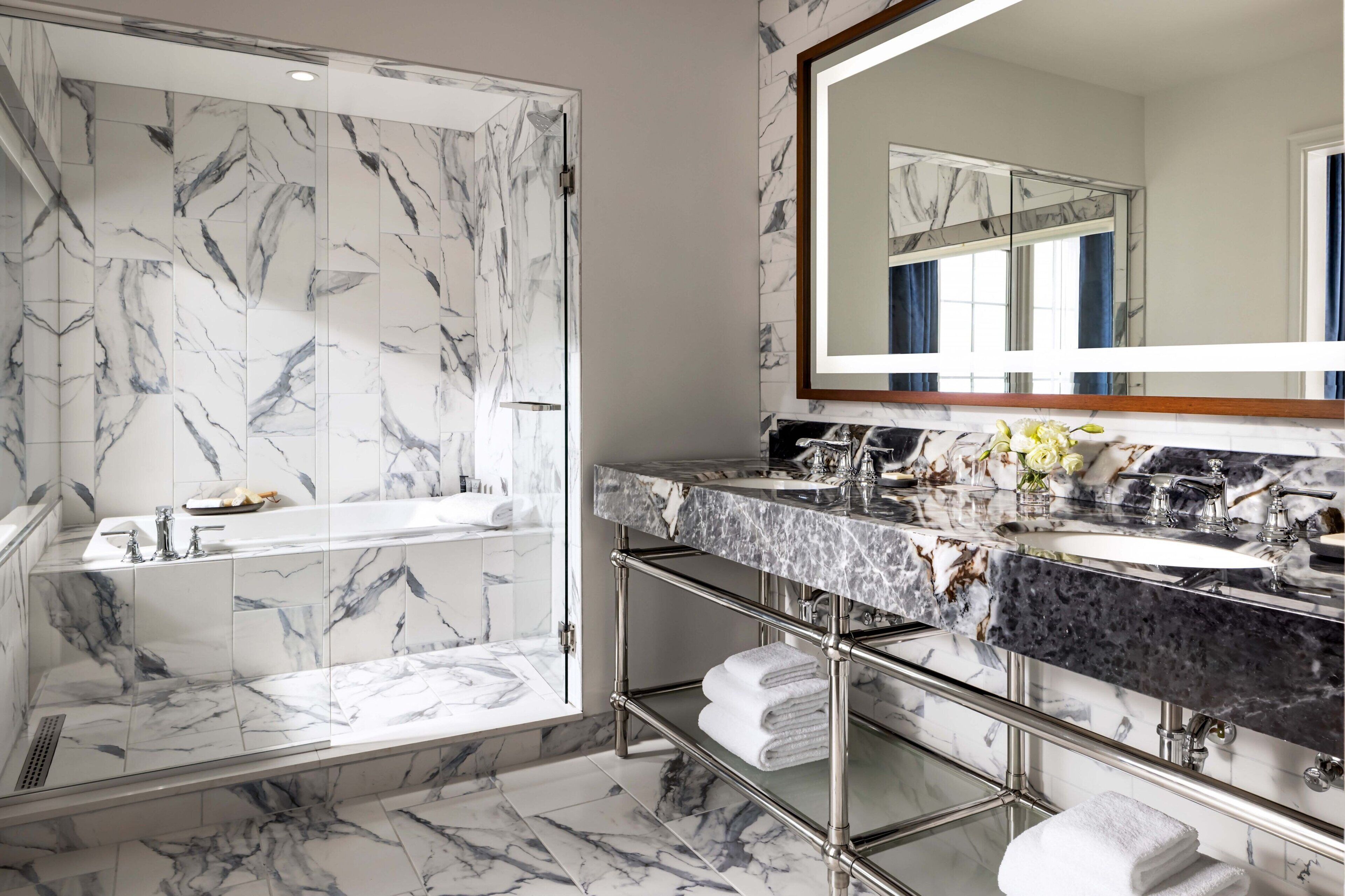 A modern bathroom with marble tiles, a glass-enclosed shower, a vanity with a large mirror, and a glass shelf.