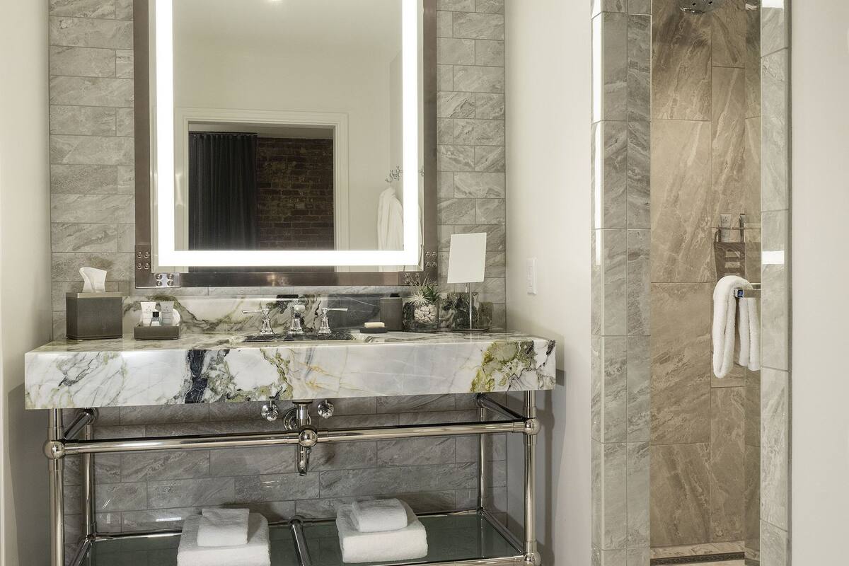 A modern bathroom with a marble vanity, a large mirror, a glass shower enclosure, and a white towel on the floor.