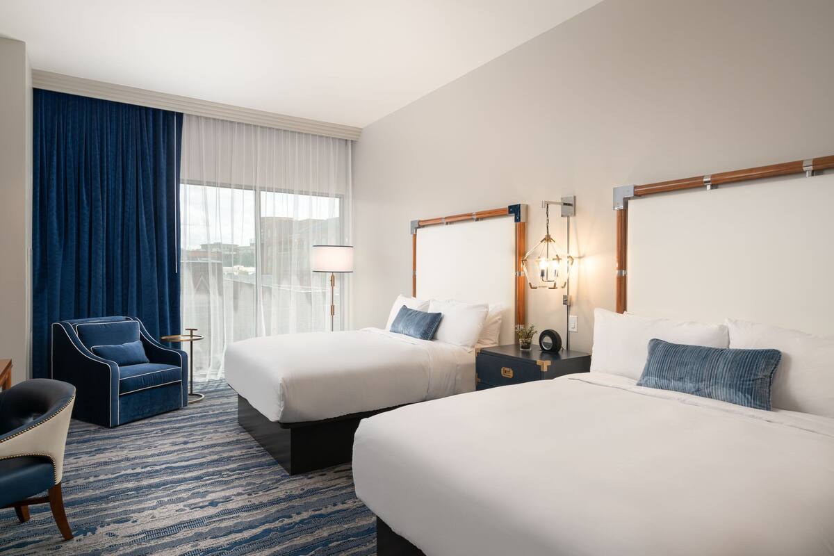 A hotel room with two beds, a blue armchair, a desk, and a large window with curtains.