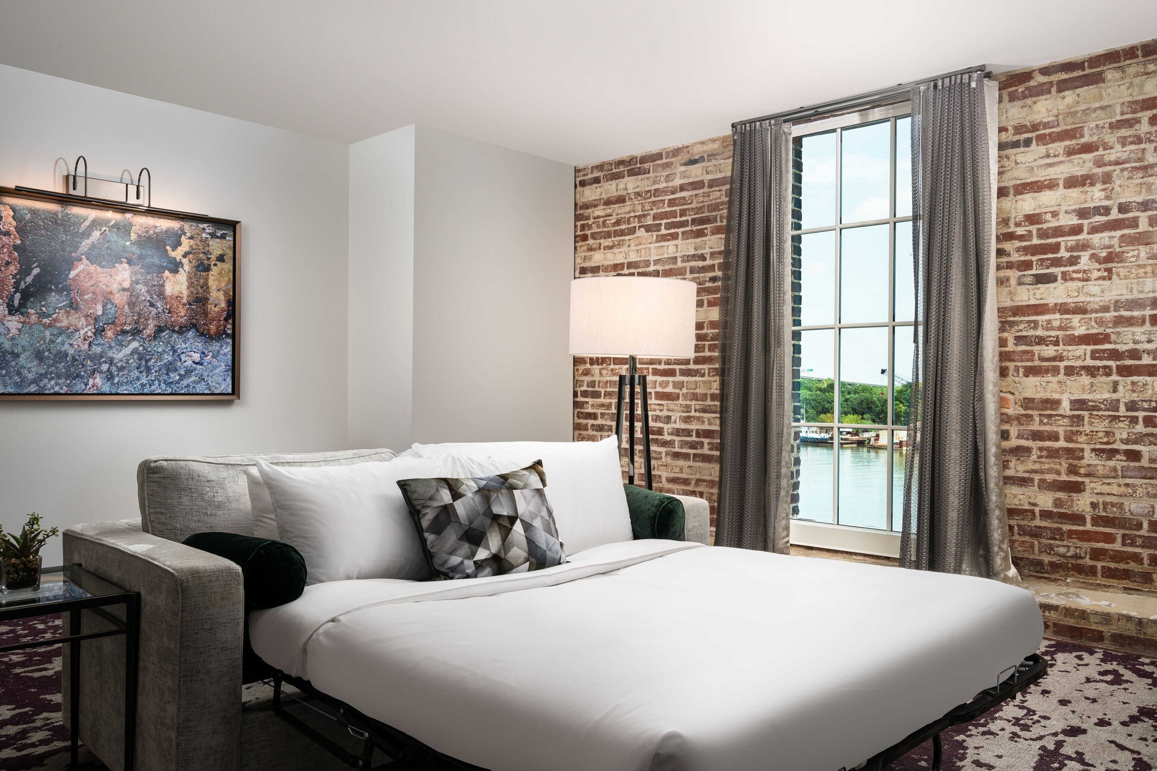 Une chambre d’hôtel avec un grand lit, un mur en briques, une fenêtre avec des rideaux, un tableau au mur et une lampe.