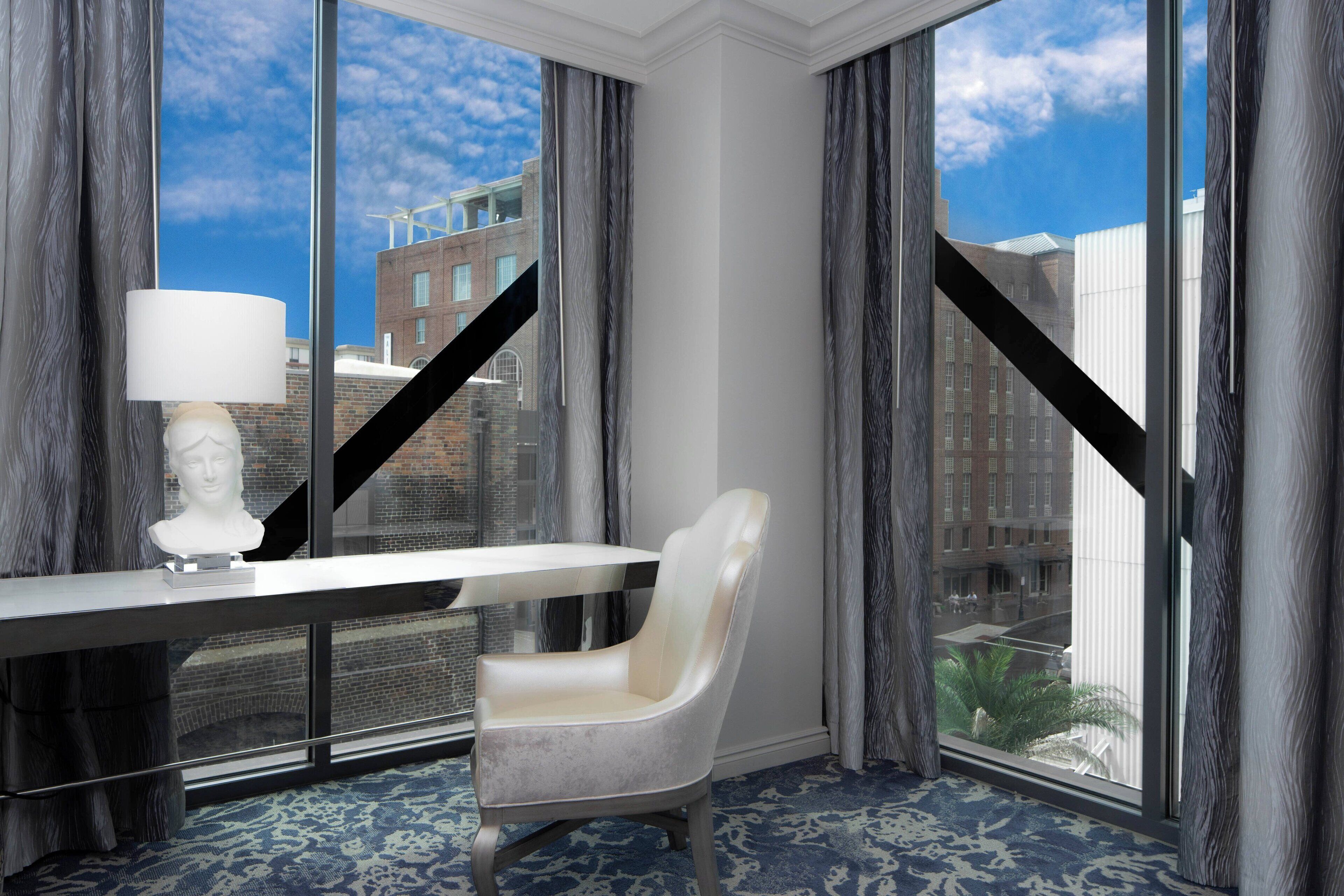 A hotel room with a large window, a white desk, a white chair, and a white lamp.