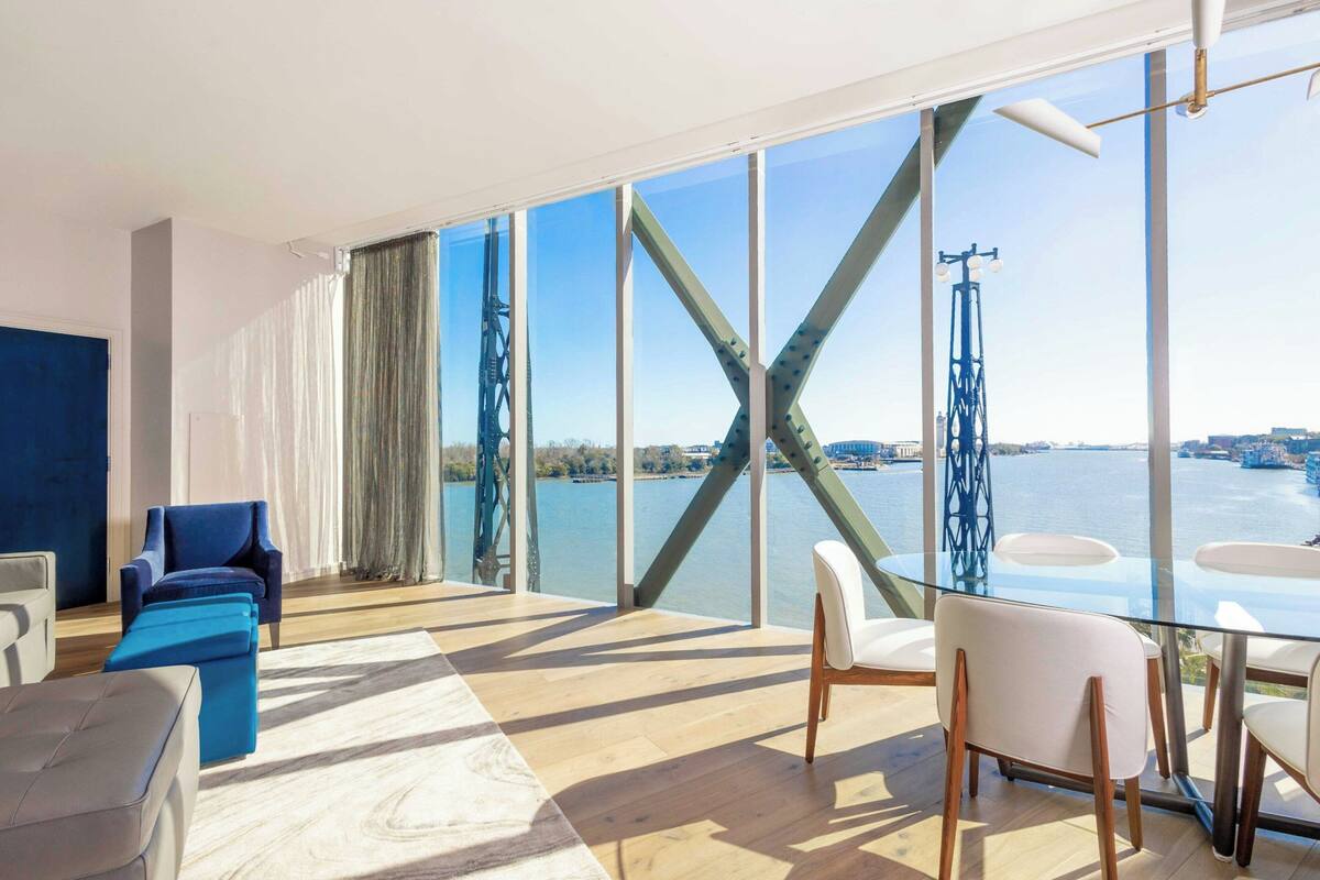 A modern living room with a large glass window, a blue sofa, a white chair, and a glass table.