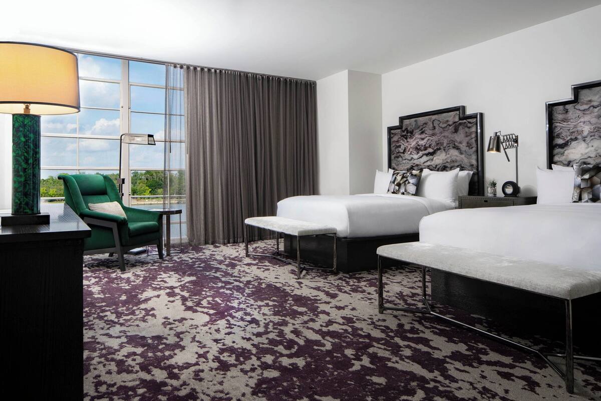 A hotel room with a large bed, a smaller bed, a green armchair, a desk, and a large window with curtains.