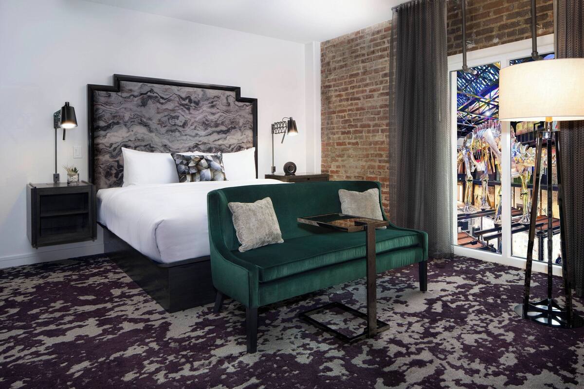 A hotel room with a bed, a green sofa, a marble headboard, a brick wall, and a view of a colorful outdoor area.