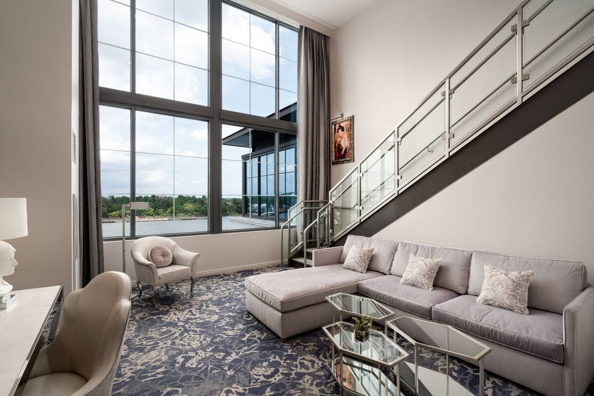 A modern hotel room with a large window, a sofa, a chair, a glass coffee table, and a staircase.