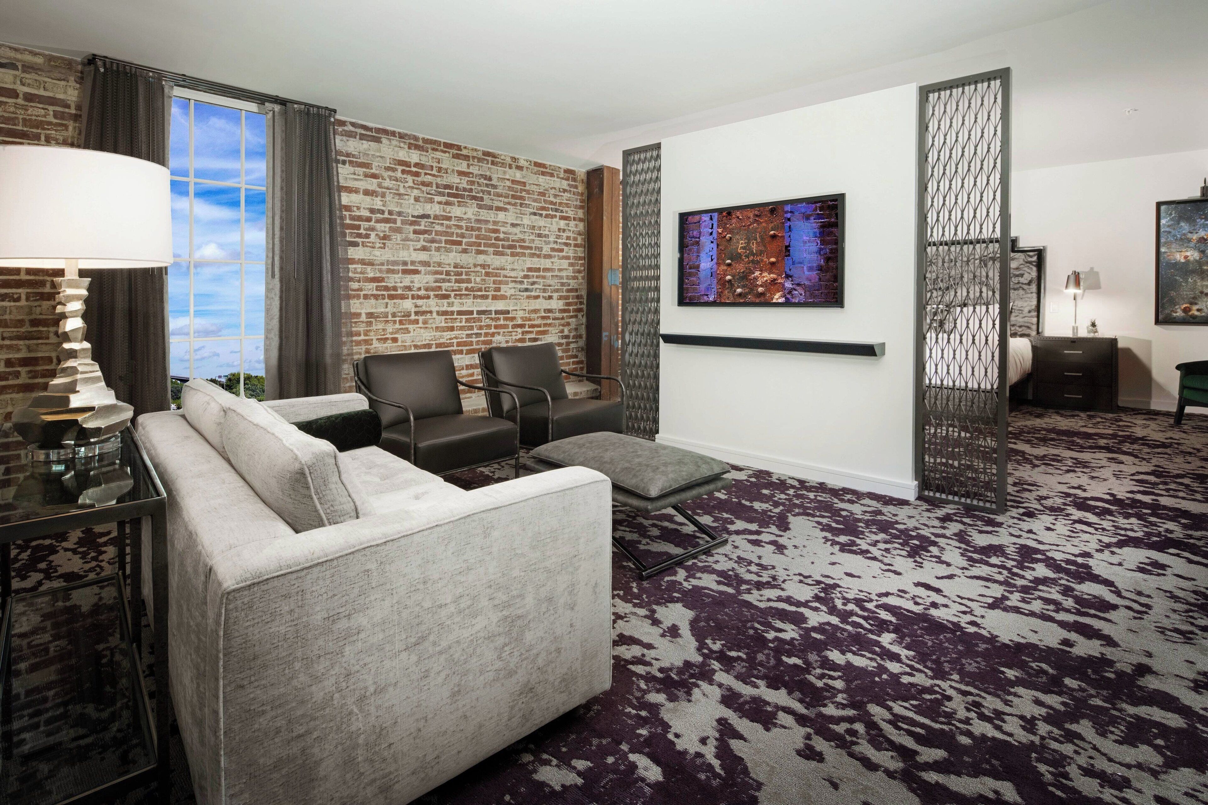 A modern hotel room with a brick wall, a large window, a gray sofa, two armchairs, a patterned carpet, and a painting on the wall.
