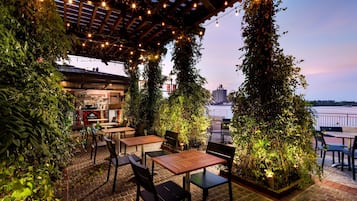 An outdoor patio with brick flooring, wooden tables and chairs, and lush greenery.