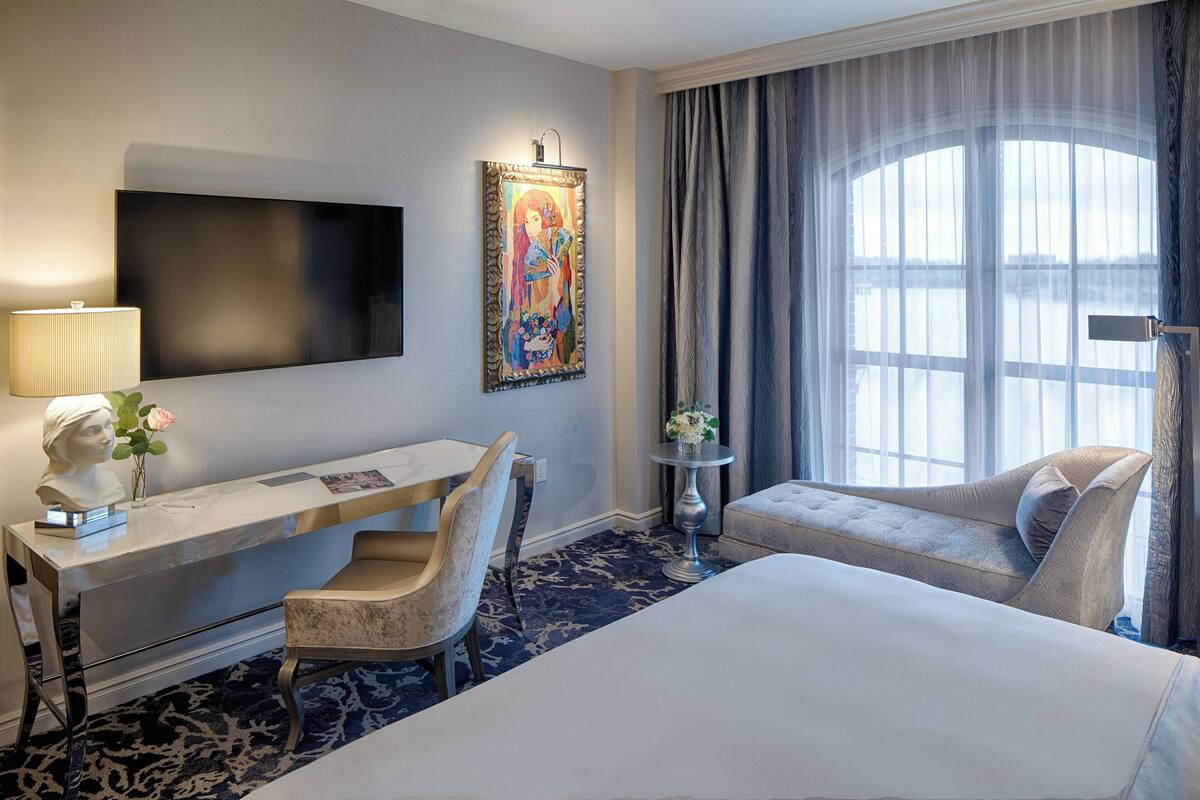 A hotel room with a bed, a desk with a chair, a TV, a painting, a window with curtains, and a small table with a vase.