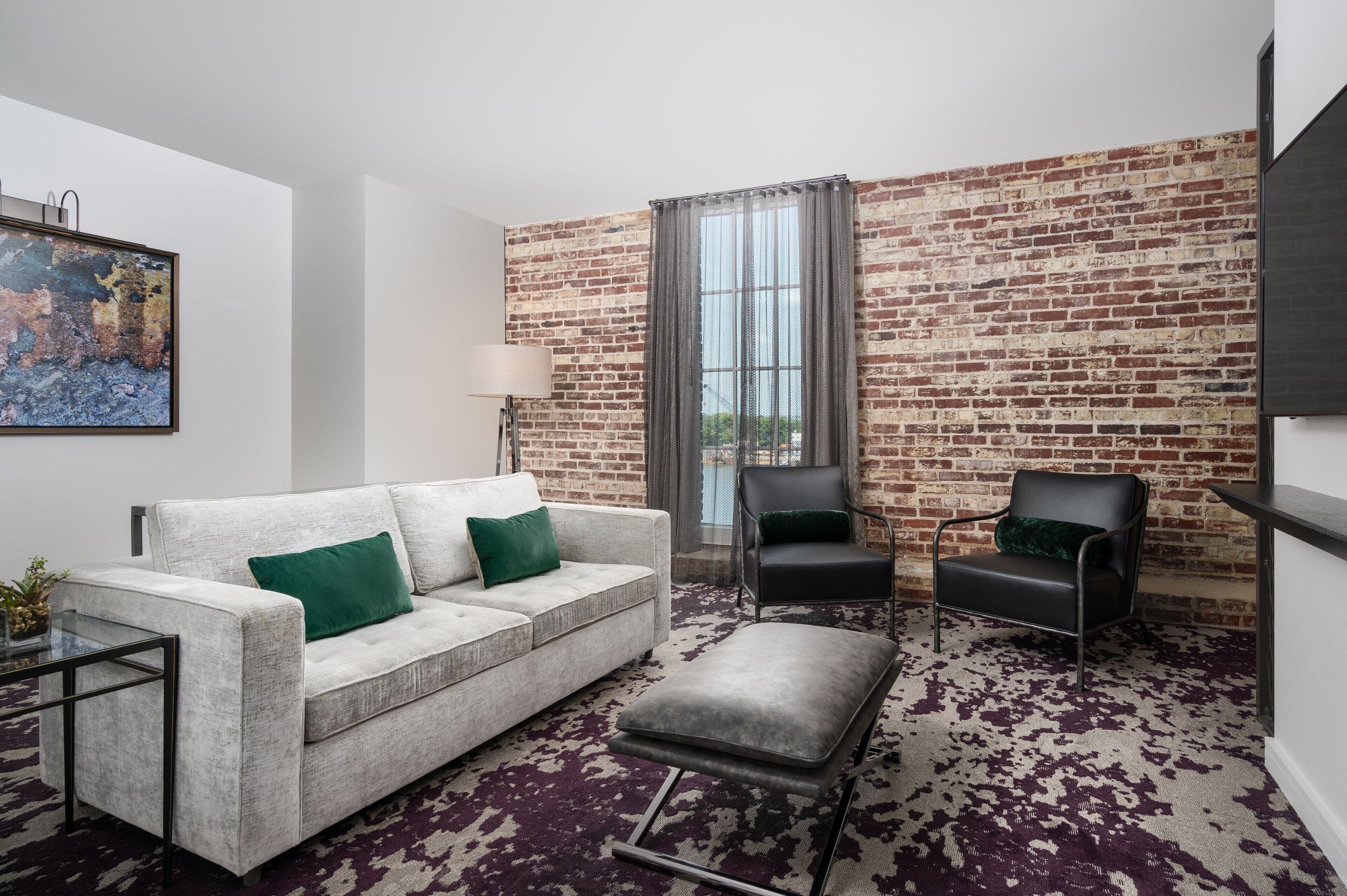 A modern living room with a white sofa, two black chairs, a glass coffee table, a patterned rug, a brick wall, and a large window with curtains.