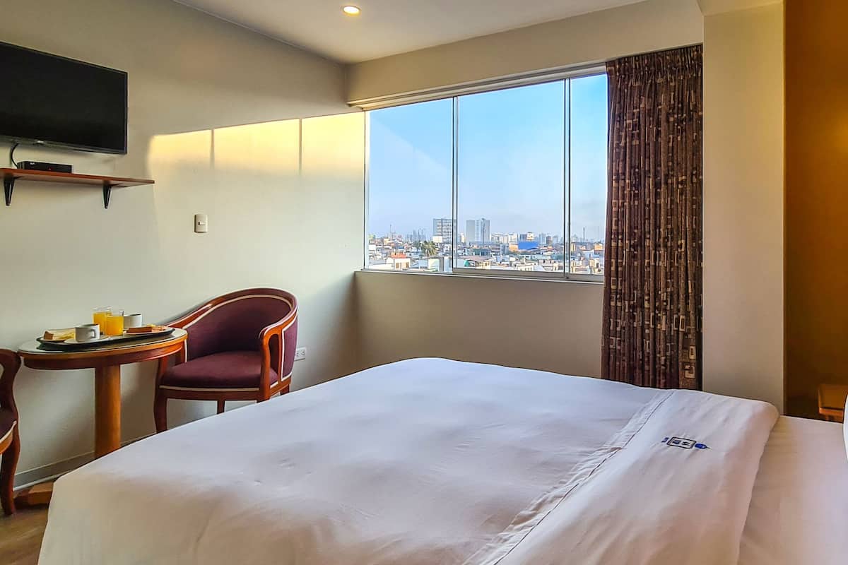 A hotel room with a bed, a chair, a small table with breakfast items, a TV, and a window with a city view.
