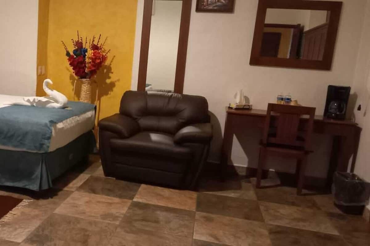 A hotel room with two beds, a brown leather chair, a small table with a vase of flowers, and a mirror on the wall.