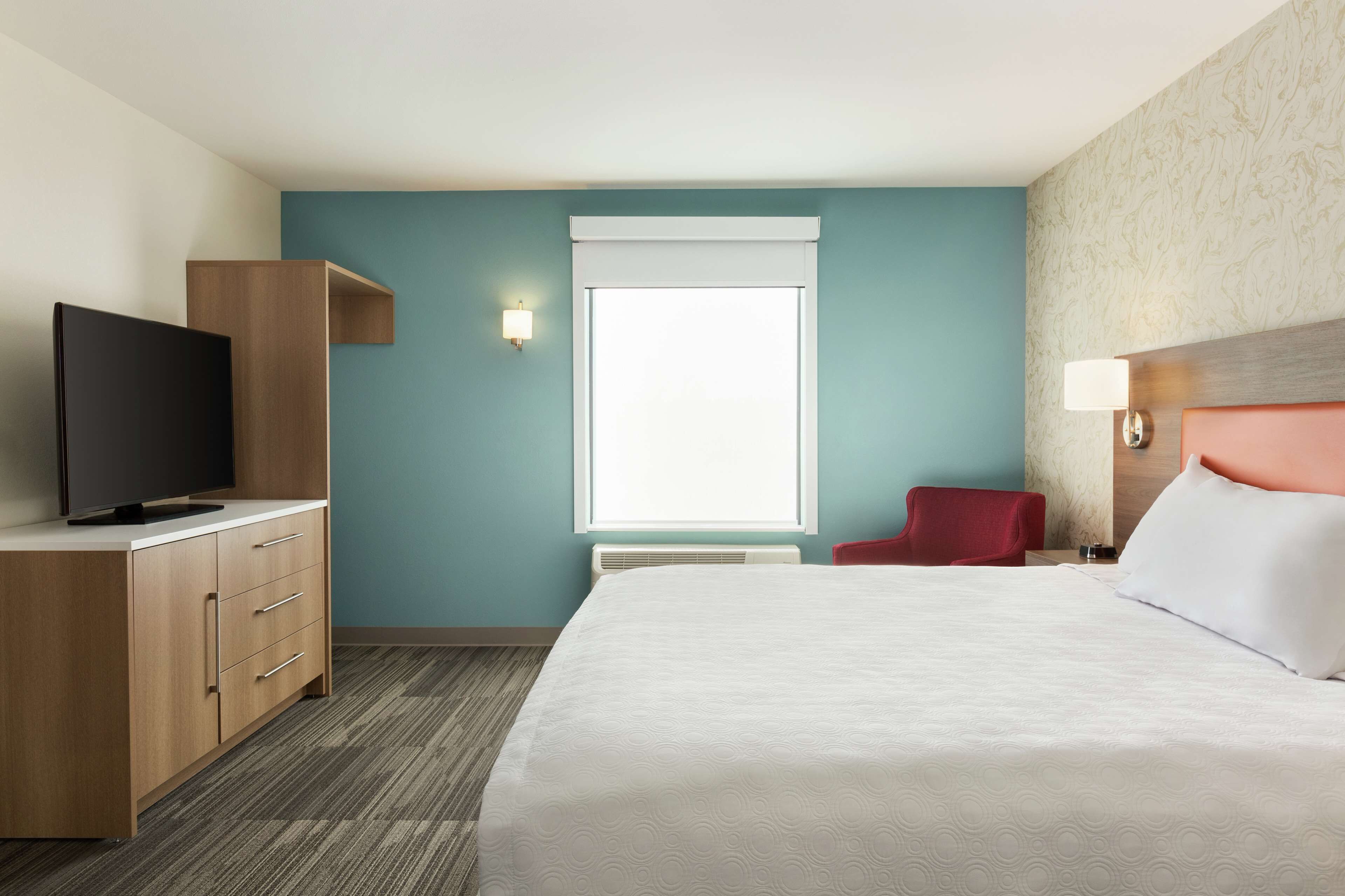 A hotel room with a bed, a TV, a red chair, a desk, and a window.