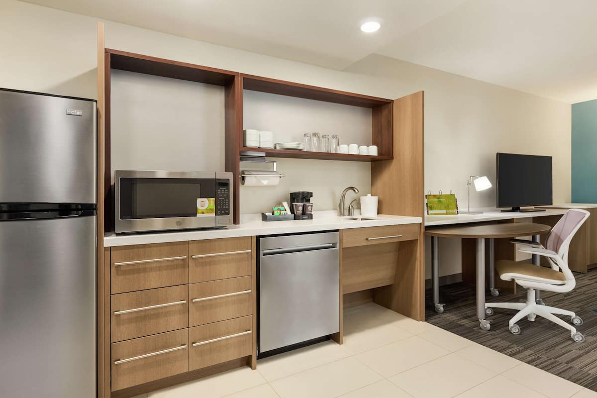Una cocina compacta con microondas integrado, refrigerador de acero inoxidable y lavavajillas. Hay una encimera de madera con un fregadero, una cafetera y una pequeña estantería con cristalería. Un escritorio con una silla está ubicado cerca del fregadero.
