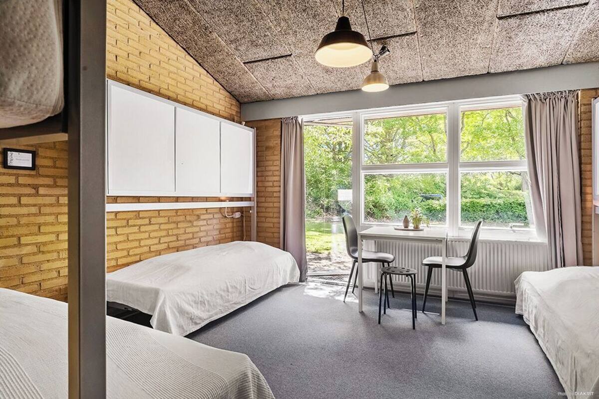 A room with two beds, a table, chairs, a radiator, and a window with a view of greenery.