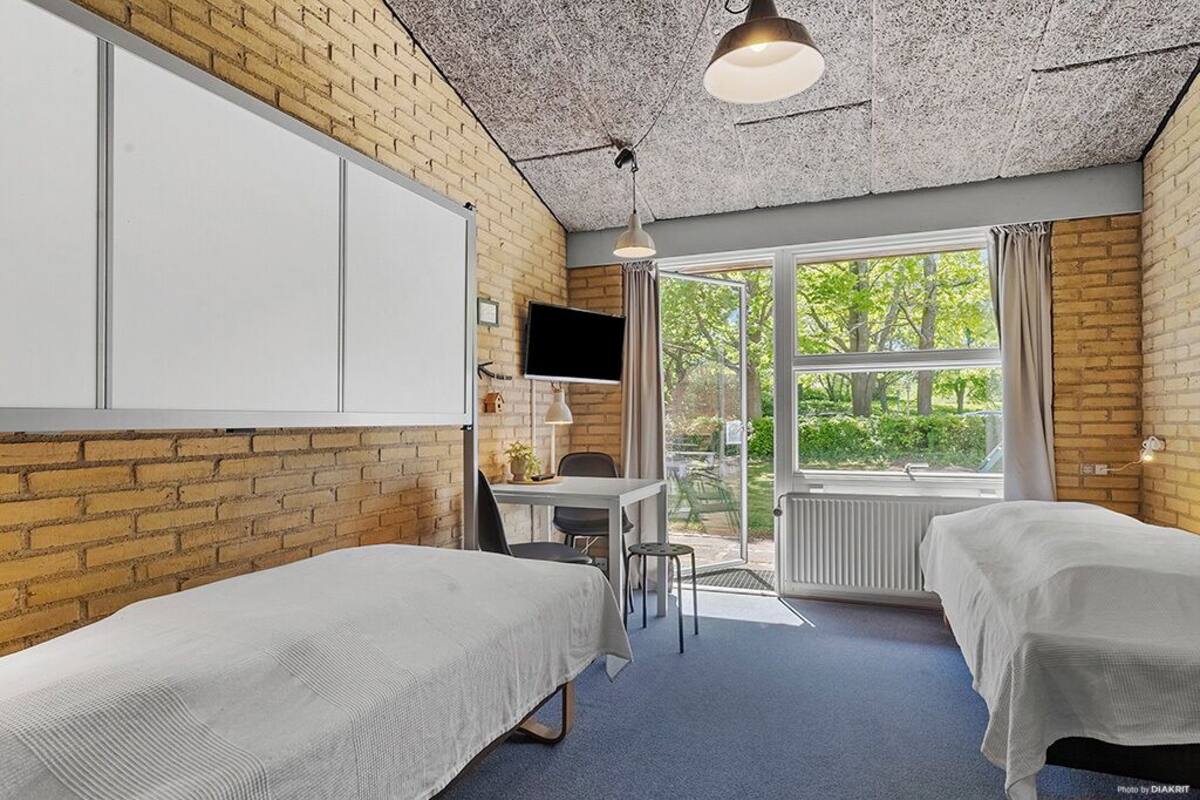 A room with two beds, a desk, a chair, a TV, and a large window with a view of greenery.