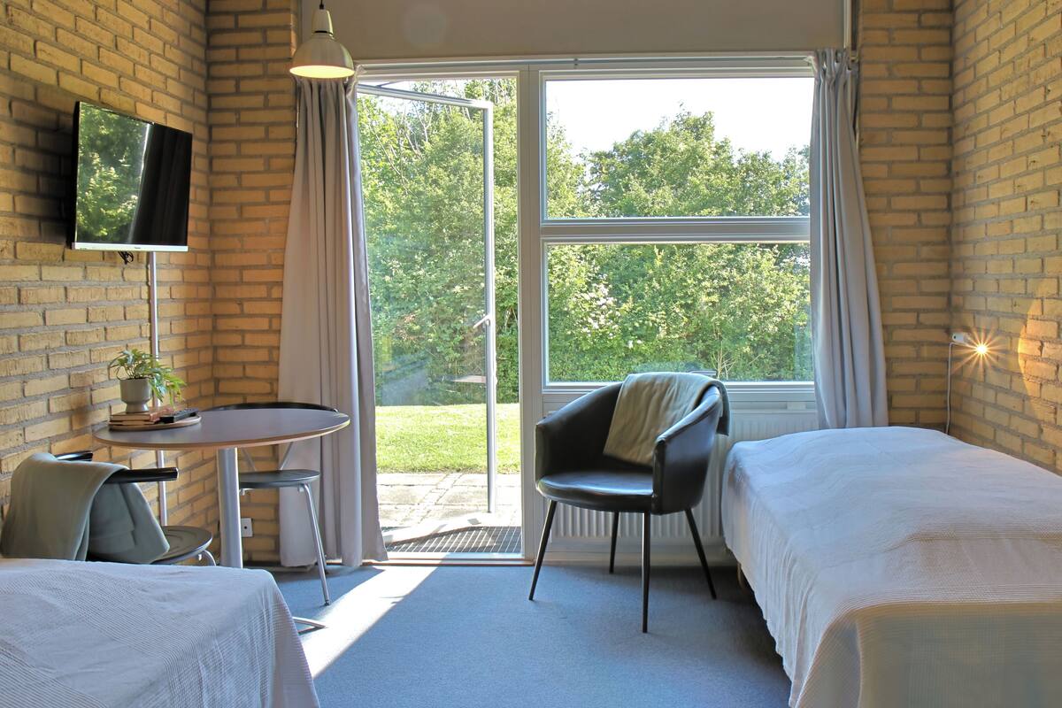 A hotel room with two beds, a TV mounted on the wall, a small table with a chair, and a large window with curtains.
