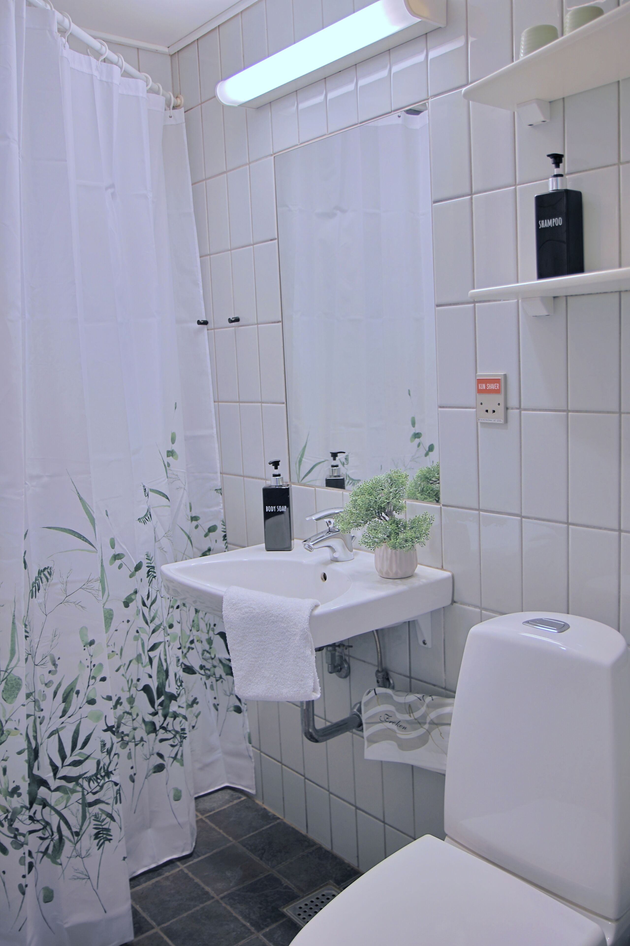 Un baño con lavamanos blanco, inodoro, cortina de ducha, estante con artículos de aseo y una planta en maceta.